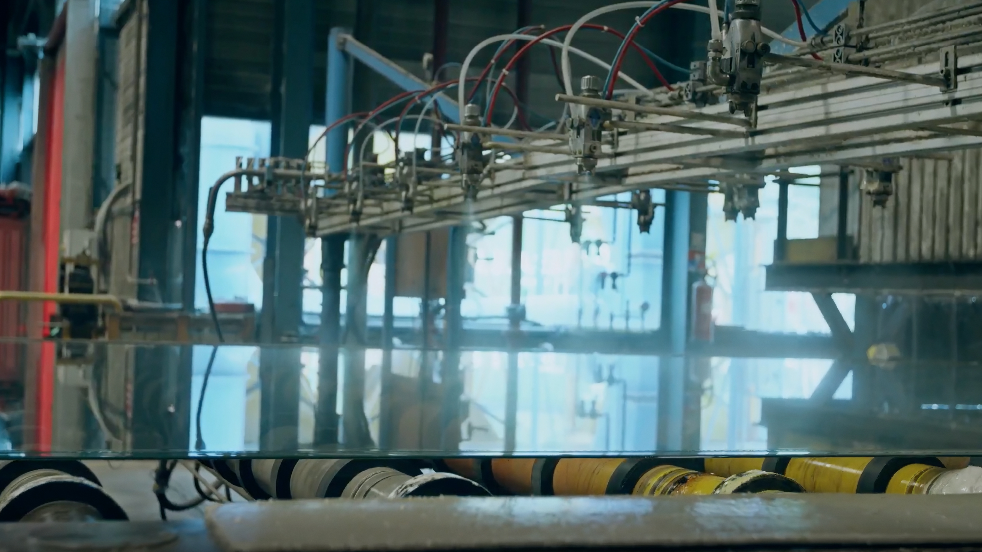 Industrial glass manufacturing equipment with overhead tubing and rollers transporting a flat sheet of glass in a factory setting.