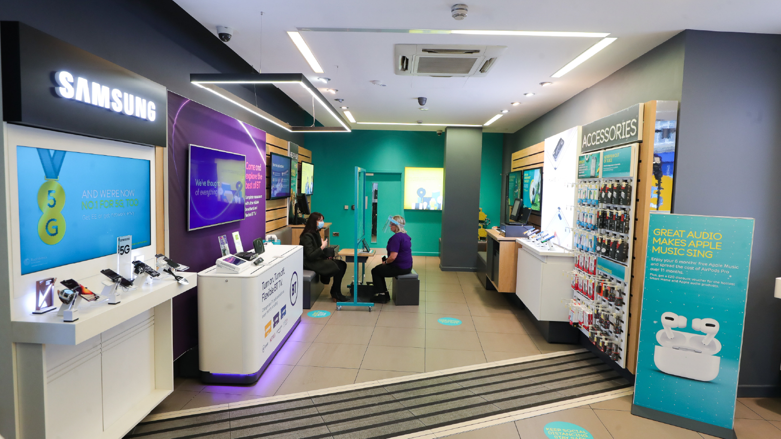 Interior of a BT retail store showing mobile devices and accessories on display, a Samsung section, service counters, and customers seated for in-store assistance.