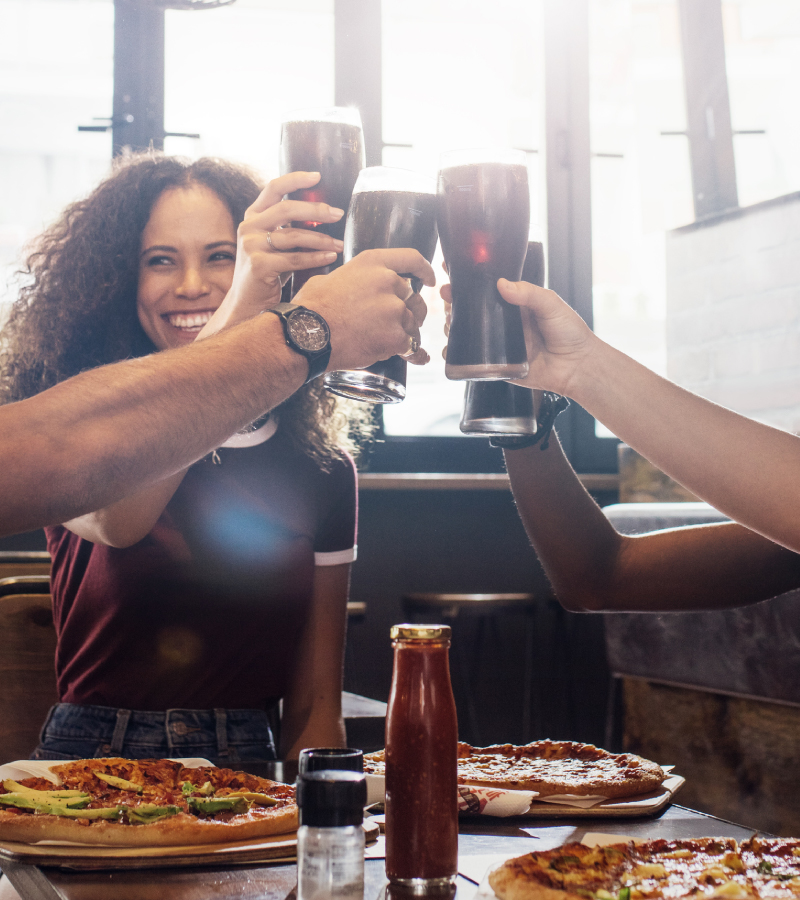A group of friends raise glasses of soda in a toast, surrounded by pizzas and condiments at a restaurant table.