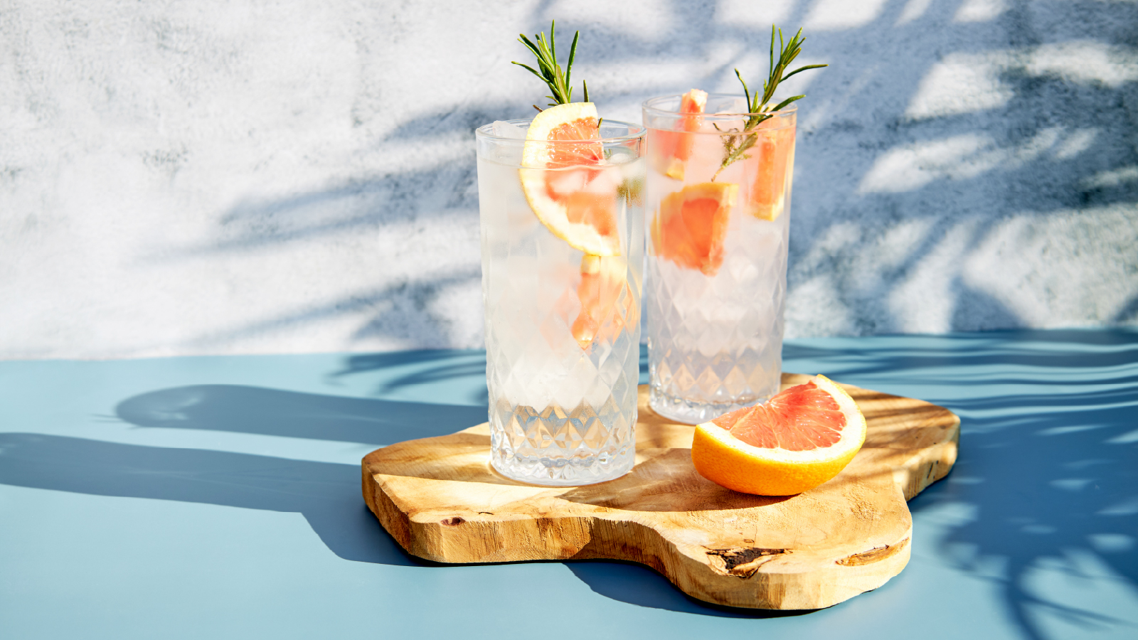 Two tall glasses of grapefruit cocktails with ice, garnished with rosemary sprigs, placed on a wooden board with a grapefruit slice.