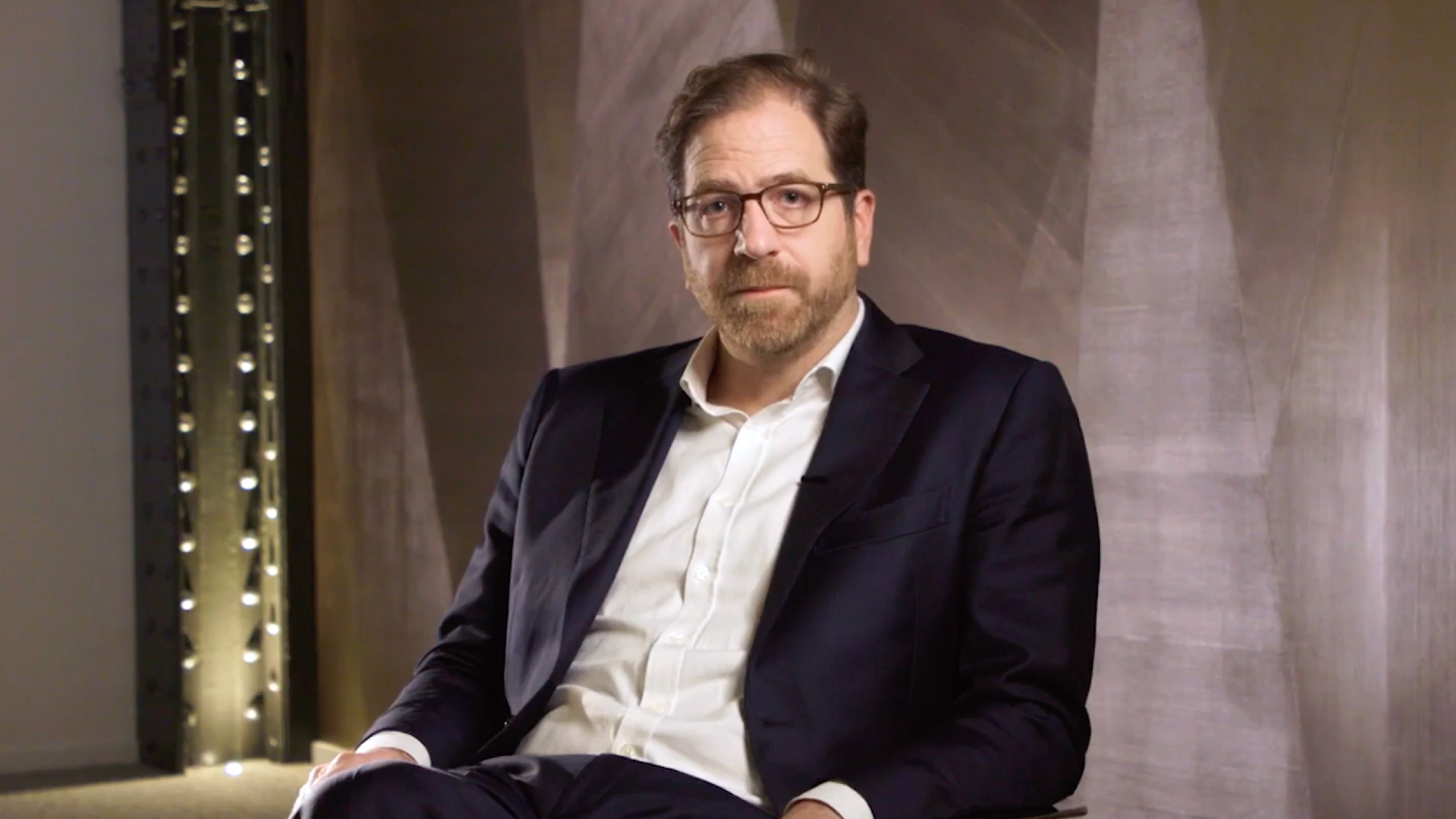 A professionally dressed man sits in a dimly lit room, looking directly at the camera during an interview or testimonial recording.