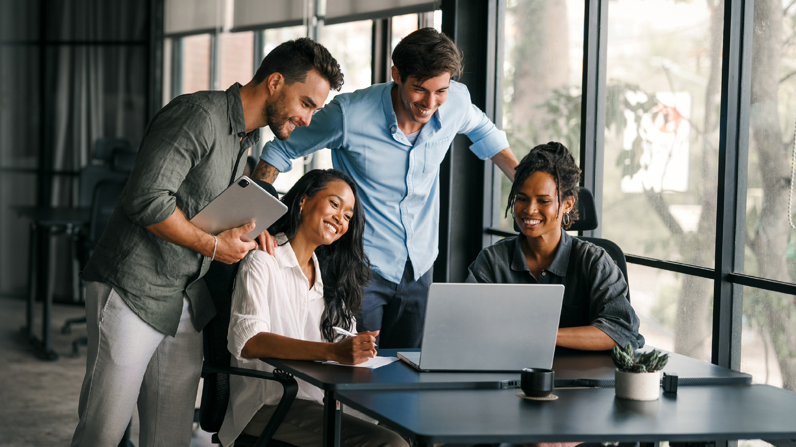 Four colleagues collaborate around a laptop in a bright modern office, smiling and reviewing work together.