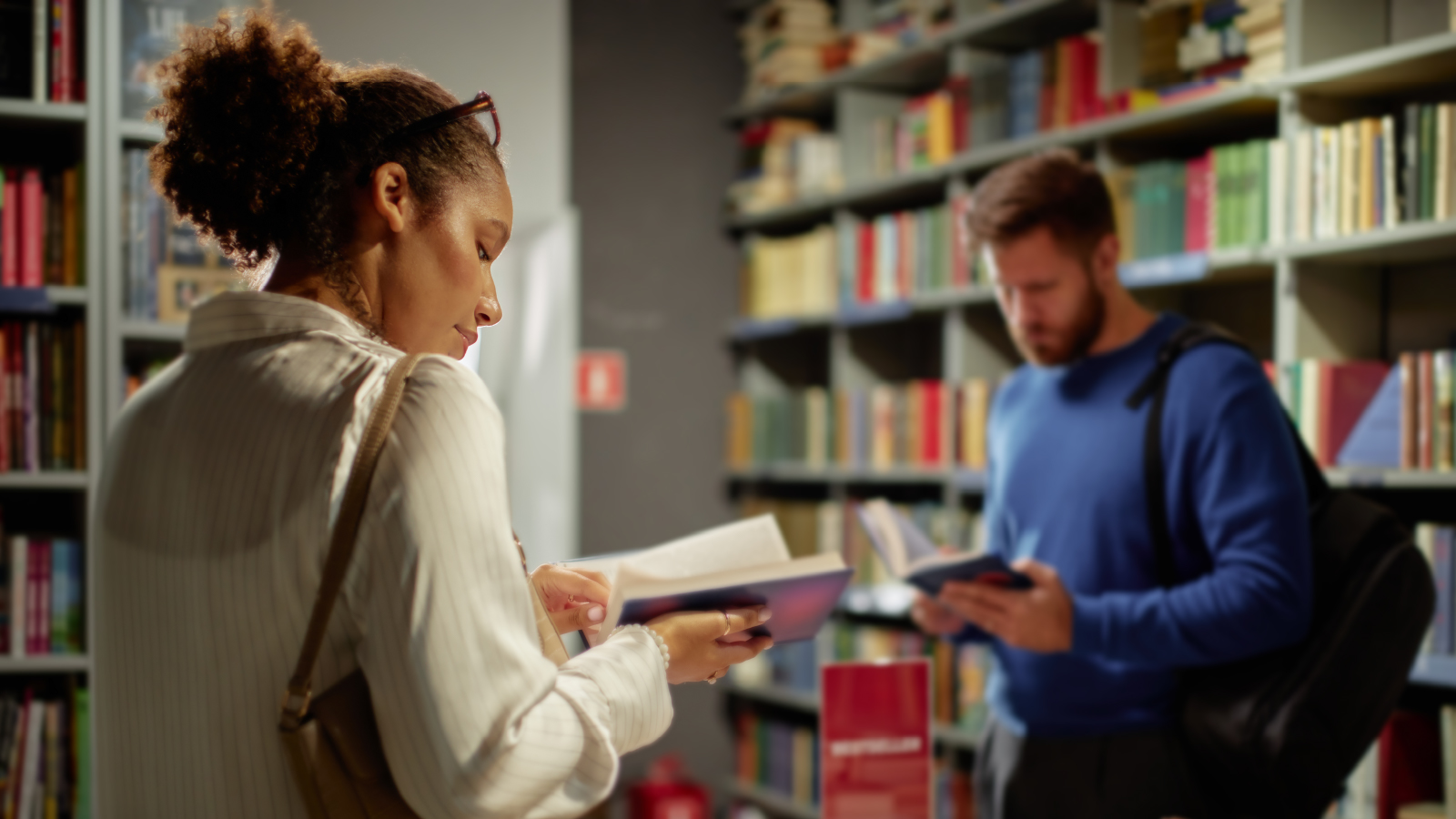 A woman in a white blouse reading a book in the foreground while a man in a blue sweater looks at another book in the background, inside a bookstore or library.