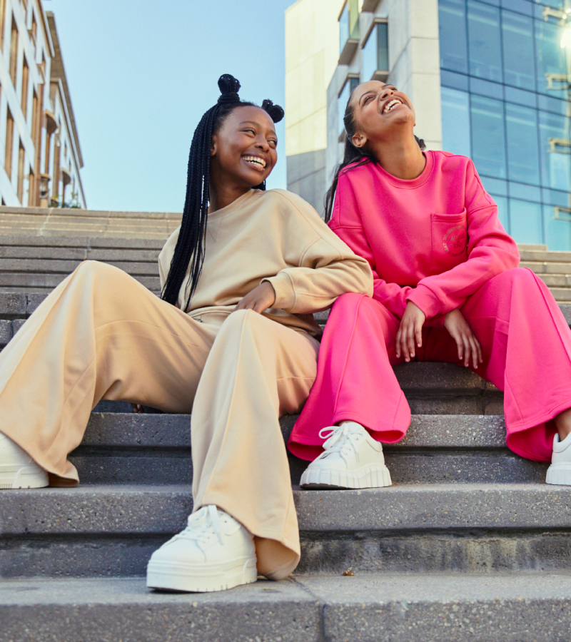 Two young women in casual outfits sitting on outdoor steps and laughing together.