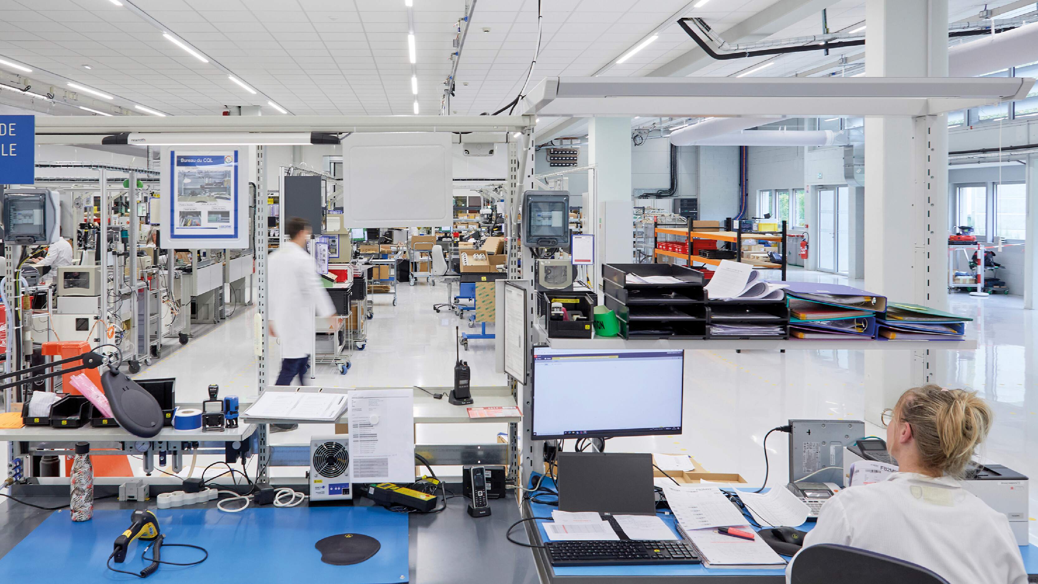 Modern manufacturing facility with employees in lab coats working at organized desks and assembly stations.