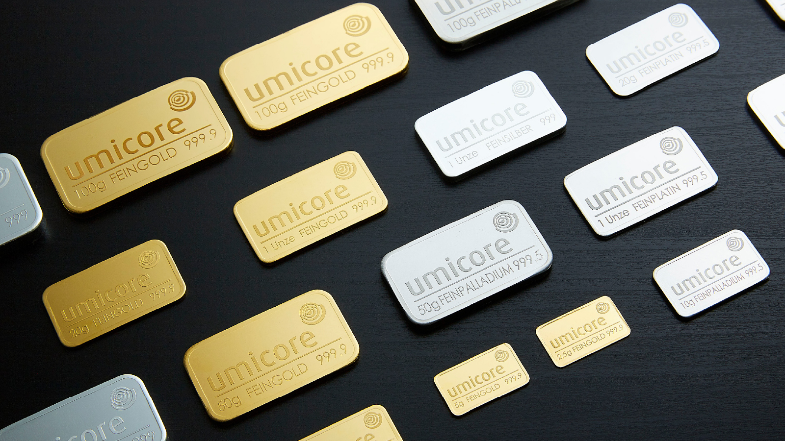 A variety of gold, silver, platinum, and palladium bars branded with the Umicore logo are arranged on a black surface, showcasing different weights and metal types.