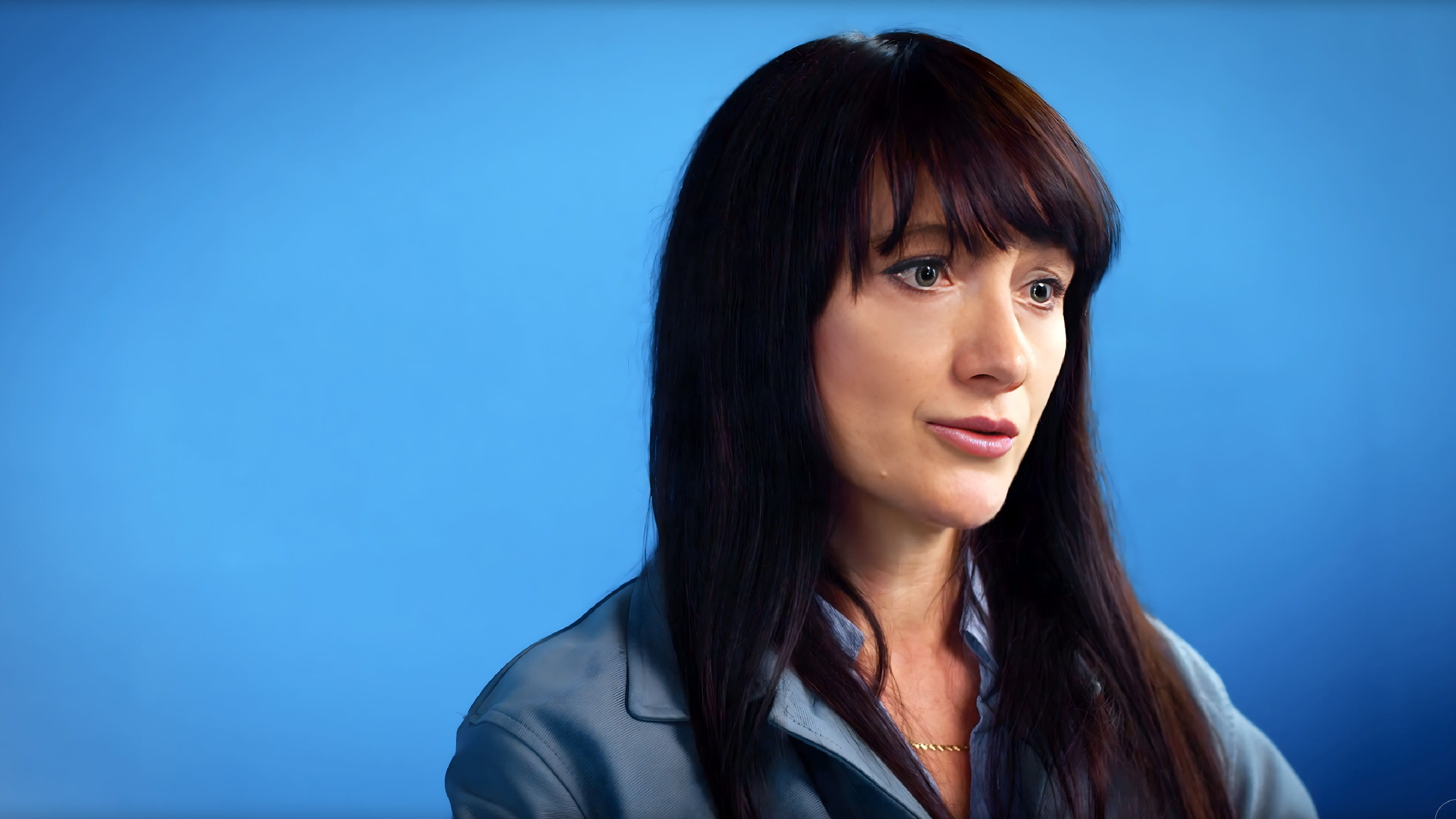 Close-up of a woman with long dark hair speaking in front of a solid blue background.