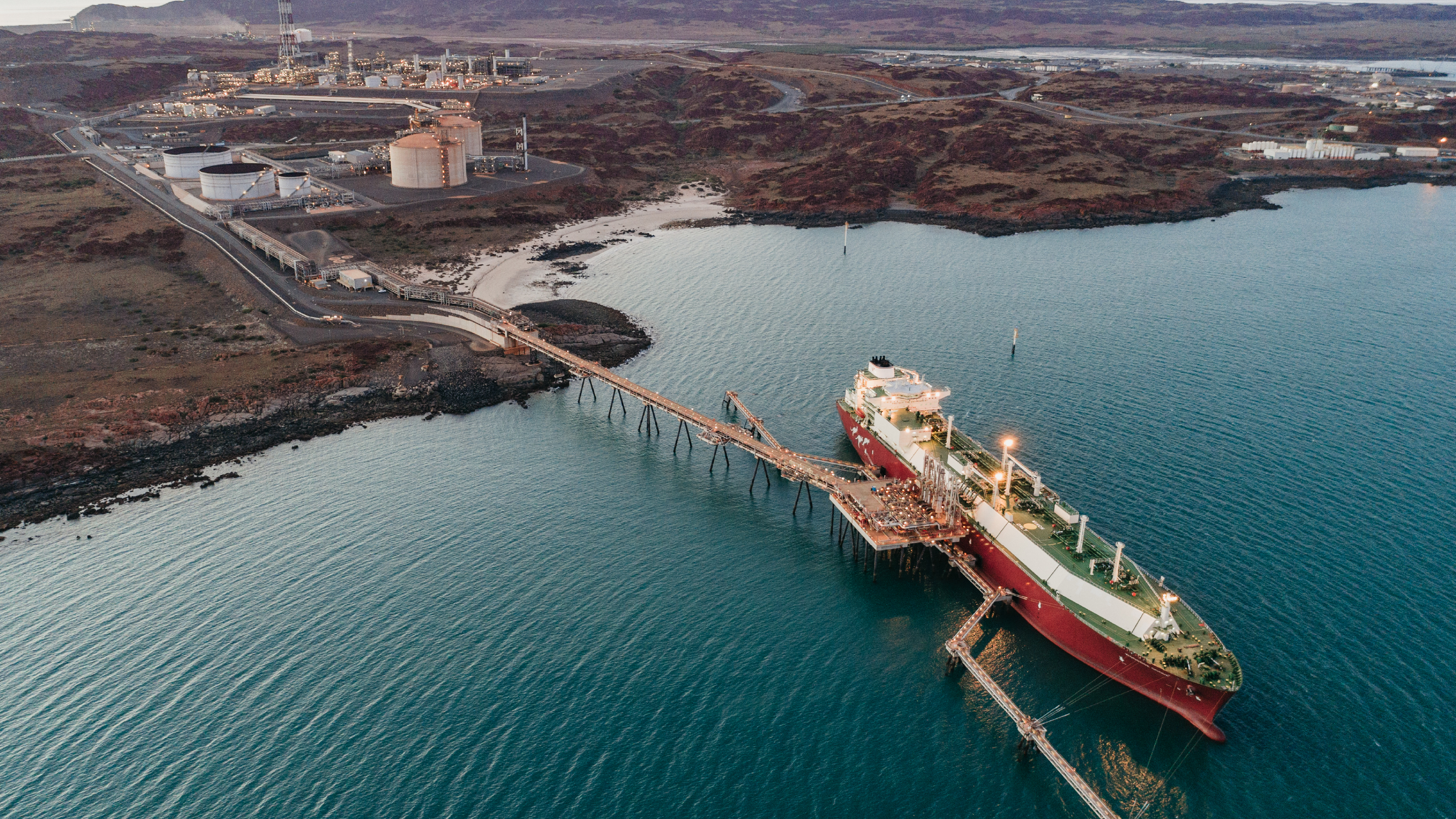 Aerial view of a coastal industrial site with storage tanks, pipelines, and a large vessel docked at a long loading pier.