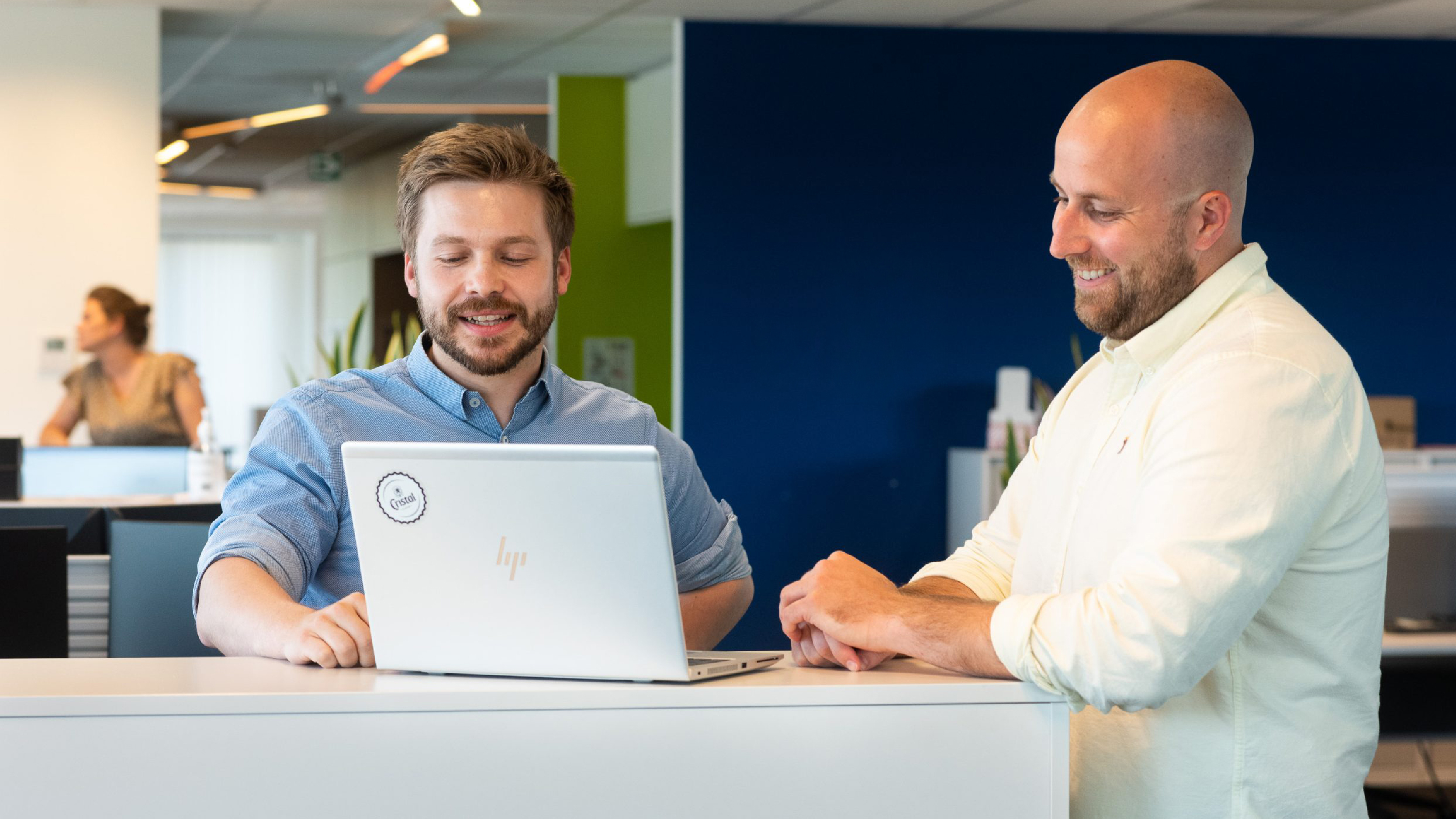 Two colleagues standing at a counter in an office environment reviewing information on a laptop, with workstations and coworkers visible in the background.