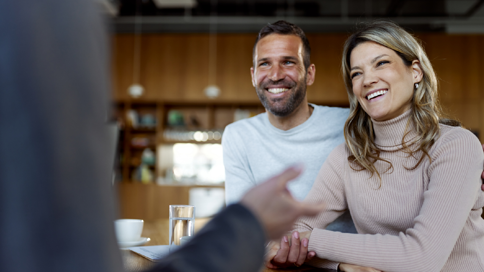 A smiling couple sits together at a table during a meeting, listening to someone who is out of frame while holding hands and leaning forward with engaged expressions.
