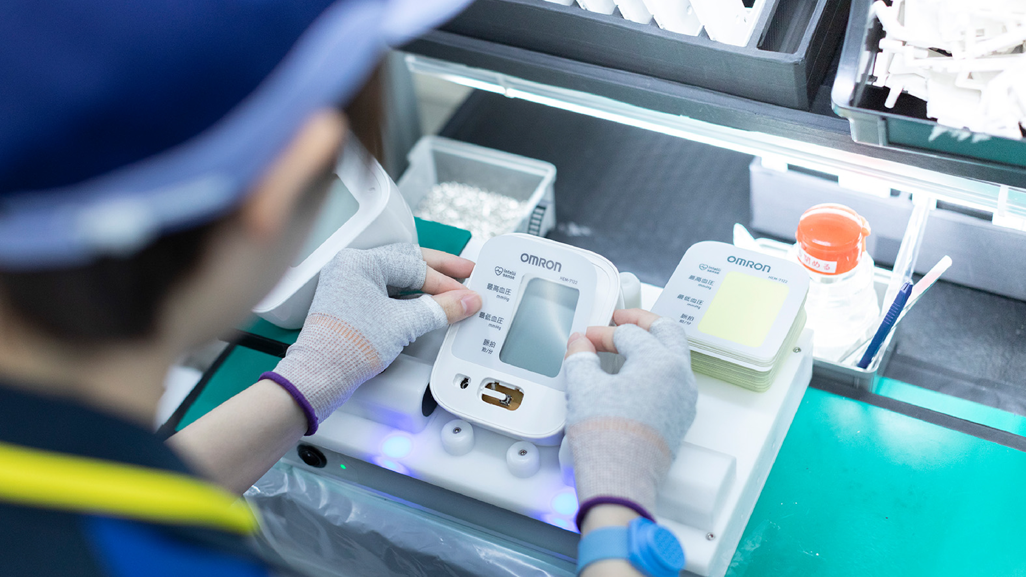 A factory worker assembling an Omron medical device, placing components onto the device housing at a workstation.
