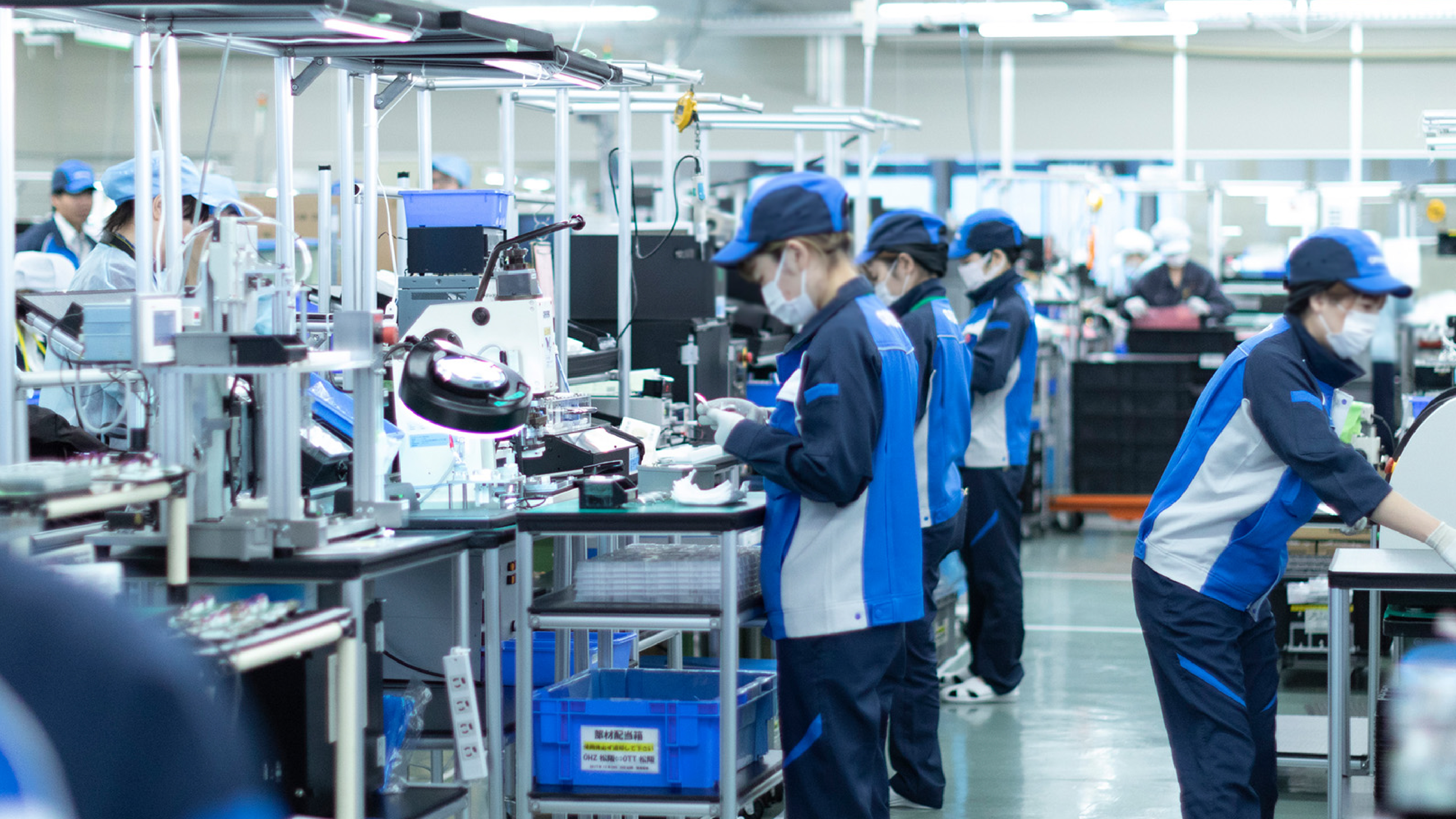 Factory workers in blue uniforms and masks operating equipment and assembling products on a busy production floor.