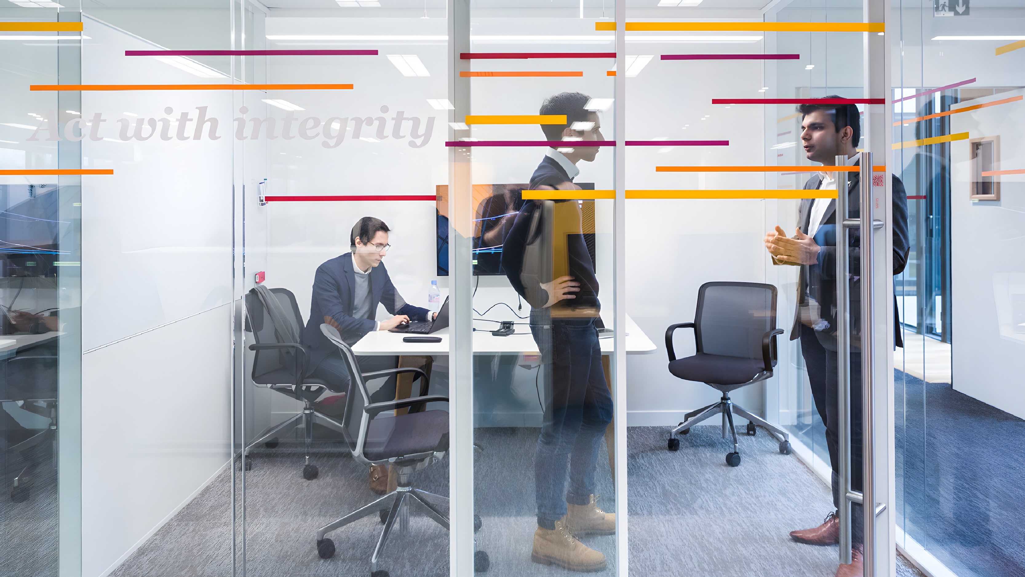 Three men inside a glass-walled office, one seated and working on a laptop while the other two stand and talk. Colored lines and the phrase "Act with integrity" are visible on the glass wall.