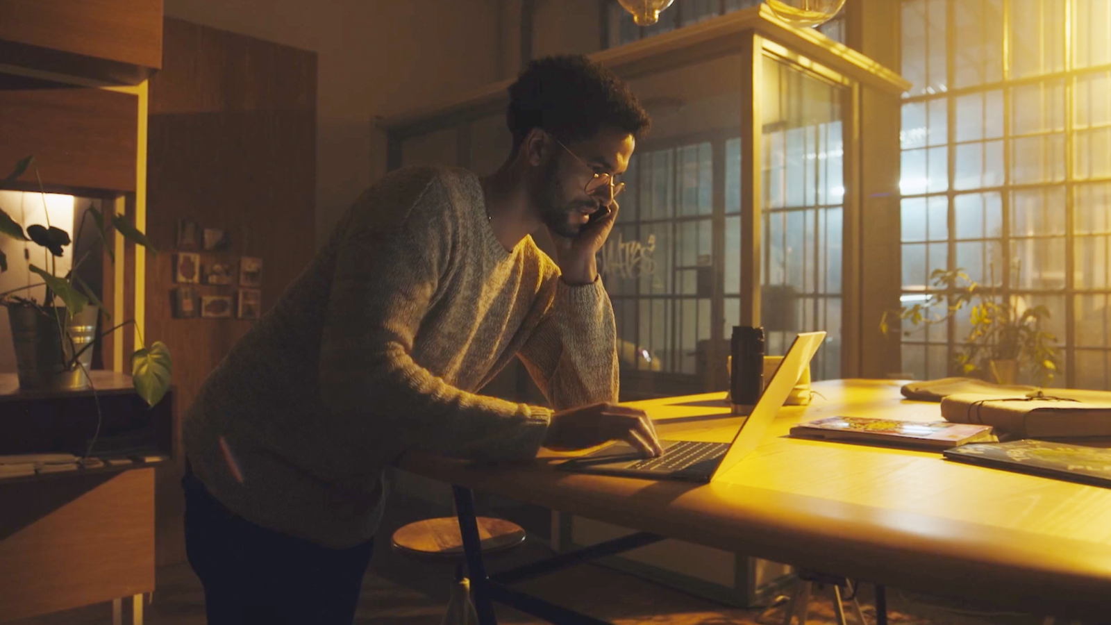 A man working late in a warmly lit office, focused on a laptop at a wooden desk.