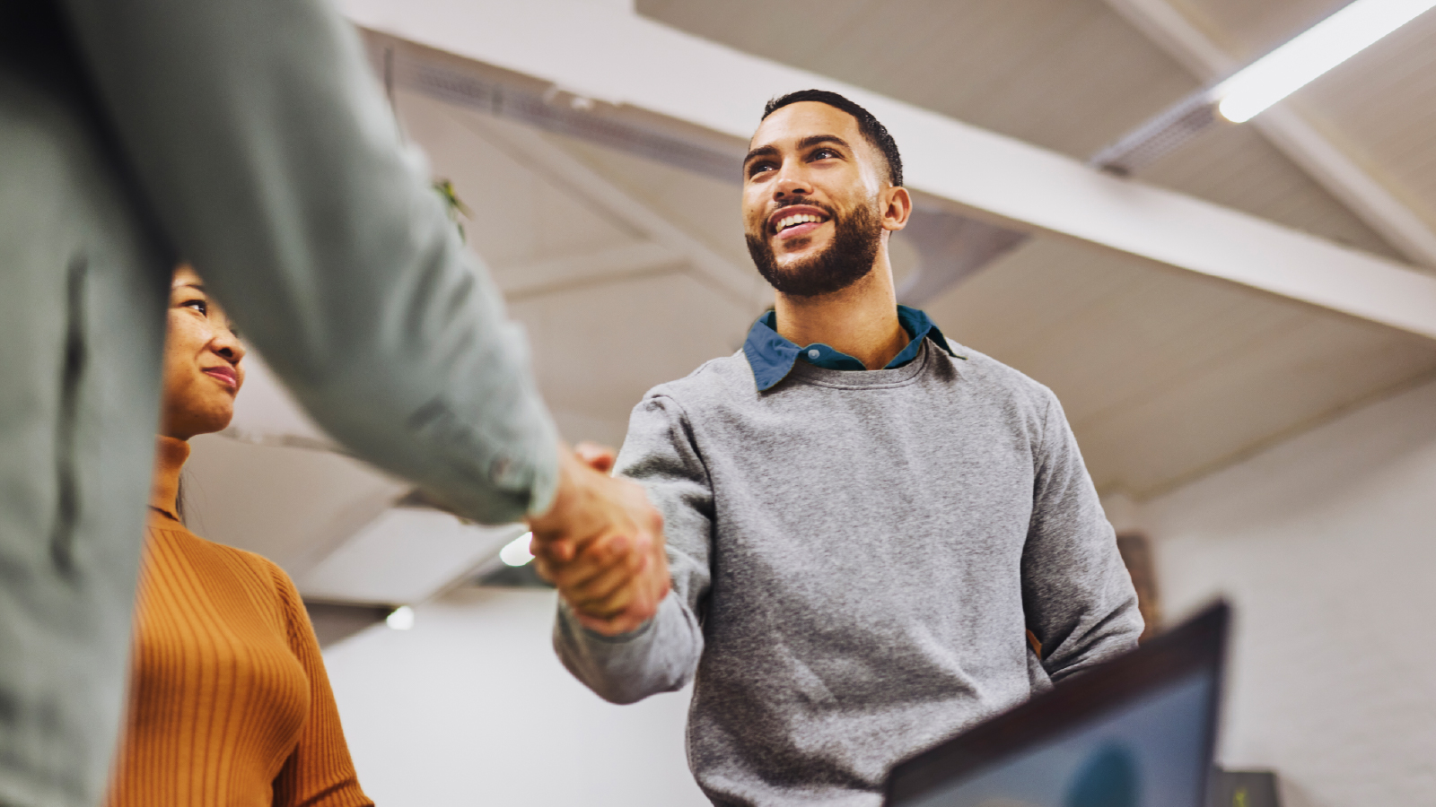 A smiling man in a gray sweater shakes hands with another person in a professional setting, with a woman in an orange sweater observing in the background. The ceiling has exposed beams and bright lighting.