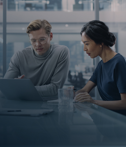 Two professionals seated at a glass table reviewing information on a laptop in a modern office, with a second scene showing two colleagues discussing and pointing at a laptop screen in a collaborative workspace.