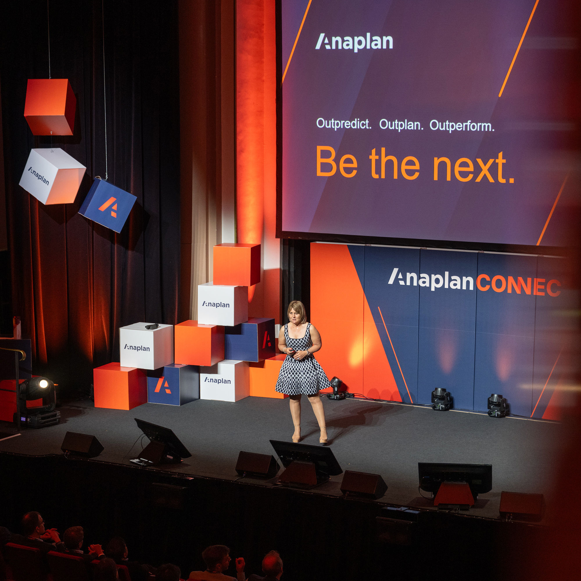 Anaplan Connect event photo. A speaker stands on stage in front of a large screen displaying the text “Outpredict. Outplan. Outperform. Be the next.” with Anaplan branding. The stage is decorated with stacked and hanging branded cubes in orange, navy, and white.