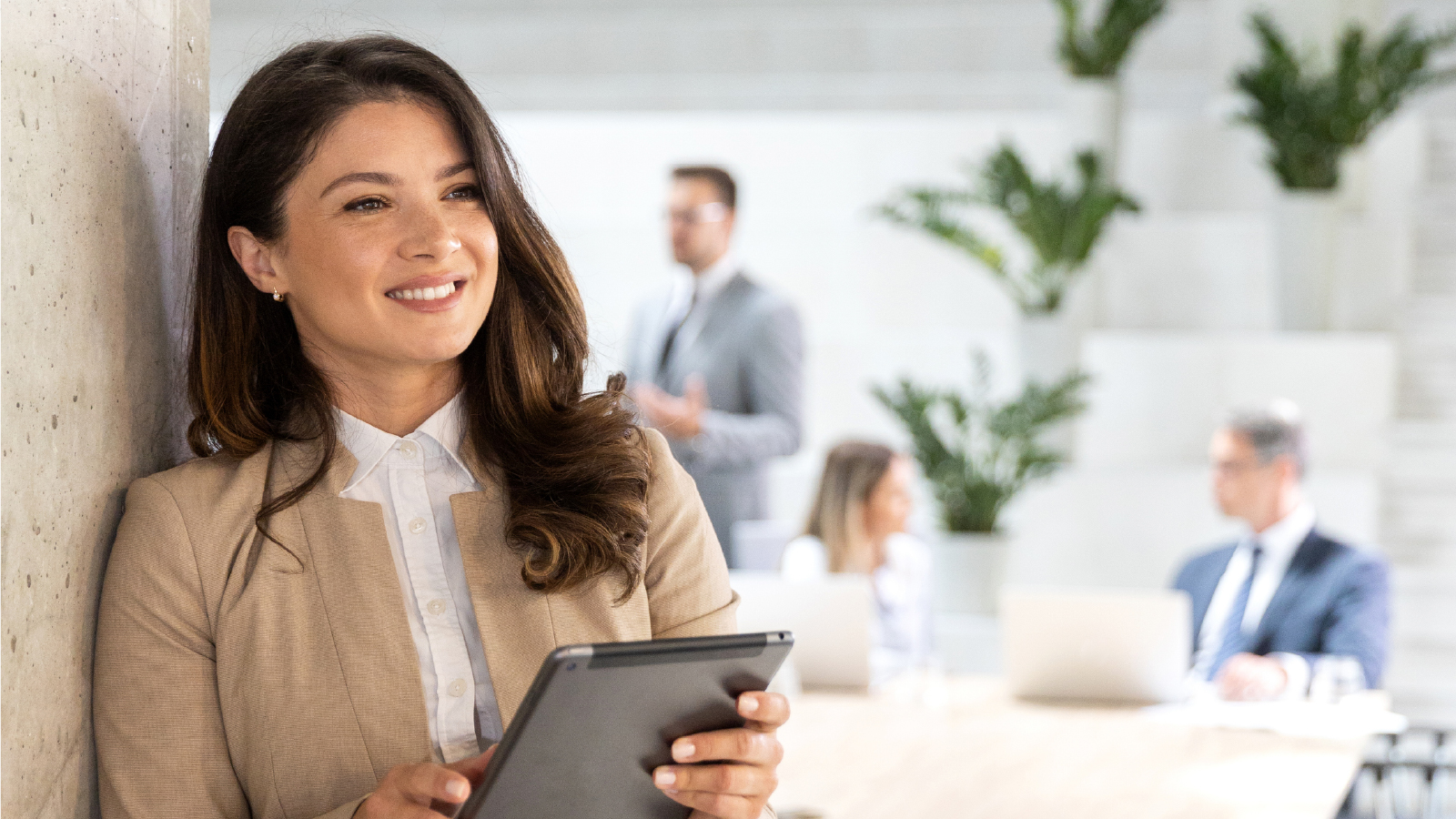 A smiling woman in a beige blazer holding a tablet, standing in a bright modern office with seated professionals and potted plants in the background.