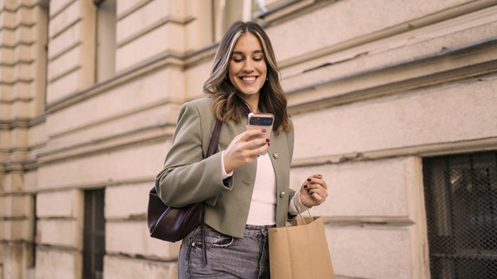 A woman walking along a city street while smiling at her phone and carrying a shopping bag, with a stone building in the background.