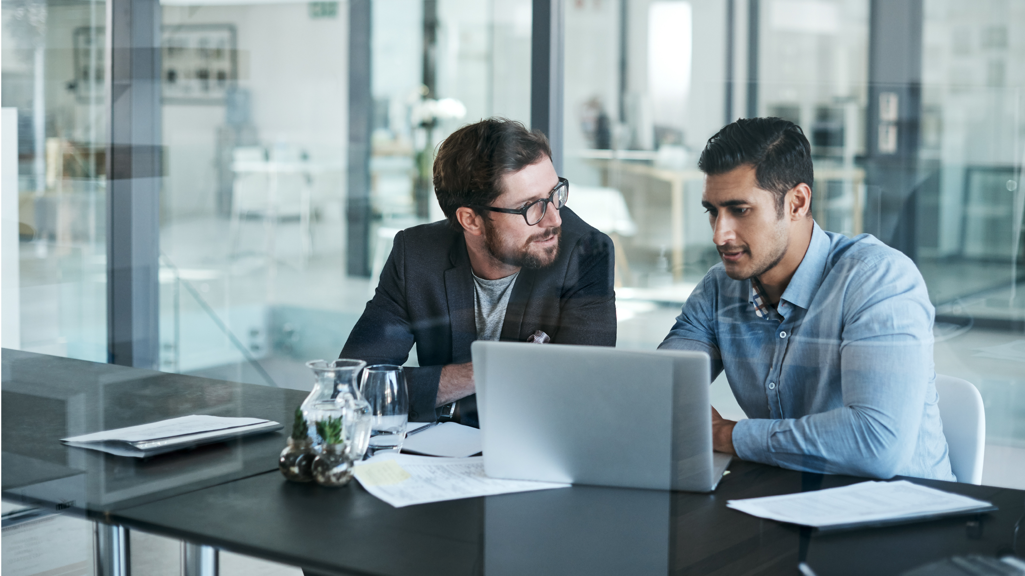 Two men seated at a glass conference table reviewing documents and a laptop, with office interiors visible in the background.