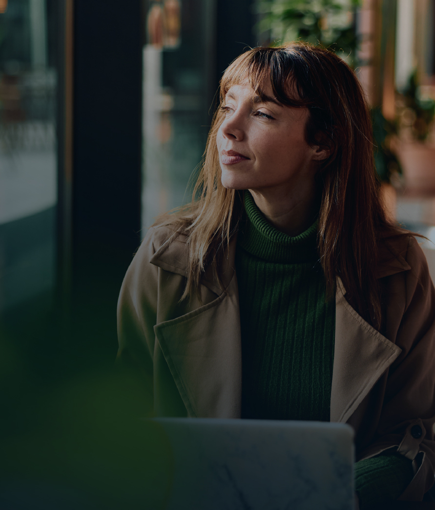 A close-up of a woman seated indoors, looking to the side while using a laptop, with a softly blurred background.