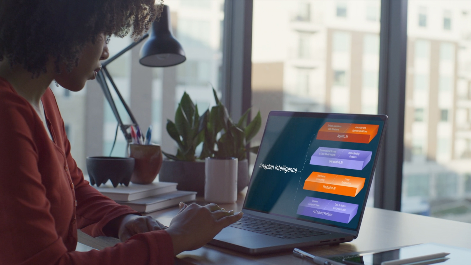 A woman sitting by a window using a laptop displaying an Anaplan Intelligence diagram with AI components.
