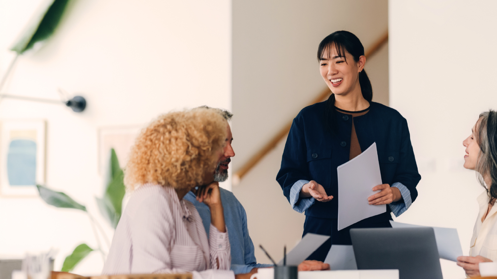 A woman in a navy blazer stands holding papers and smiling, speaking to three colleagues in a well-lit, casual meeting area with soft decor and indoor plants.