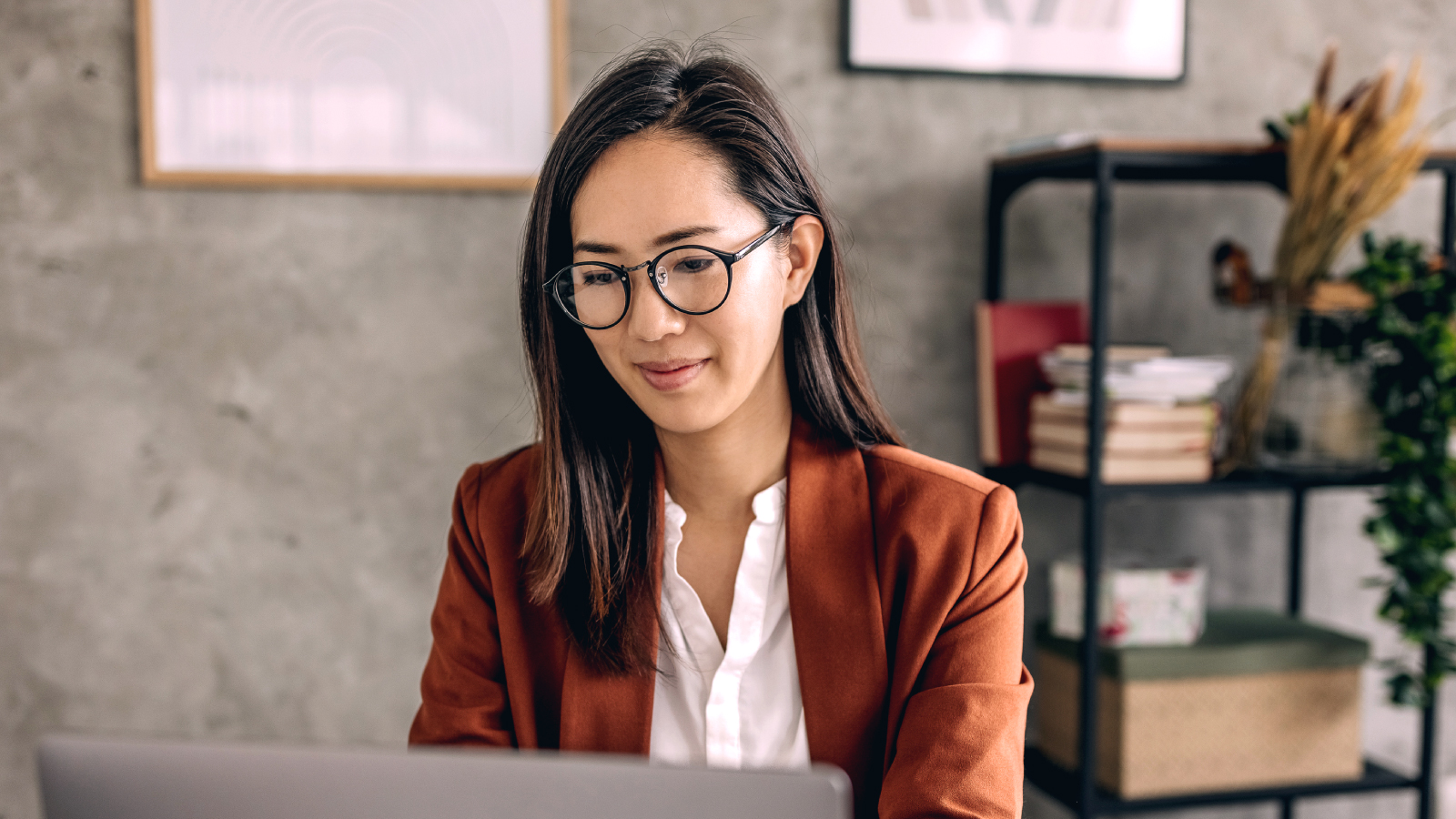 Professional woman wearing glasses and a rust-colored blazer, working on a laptop in a modern office setting.
