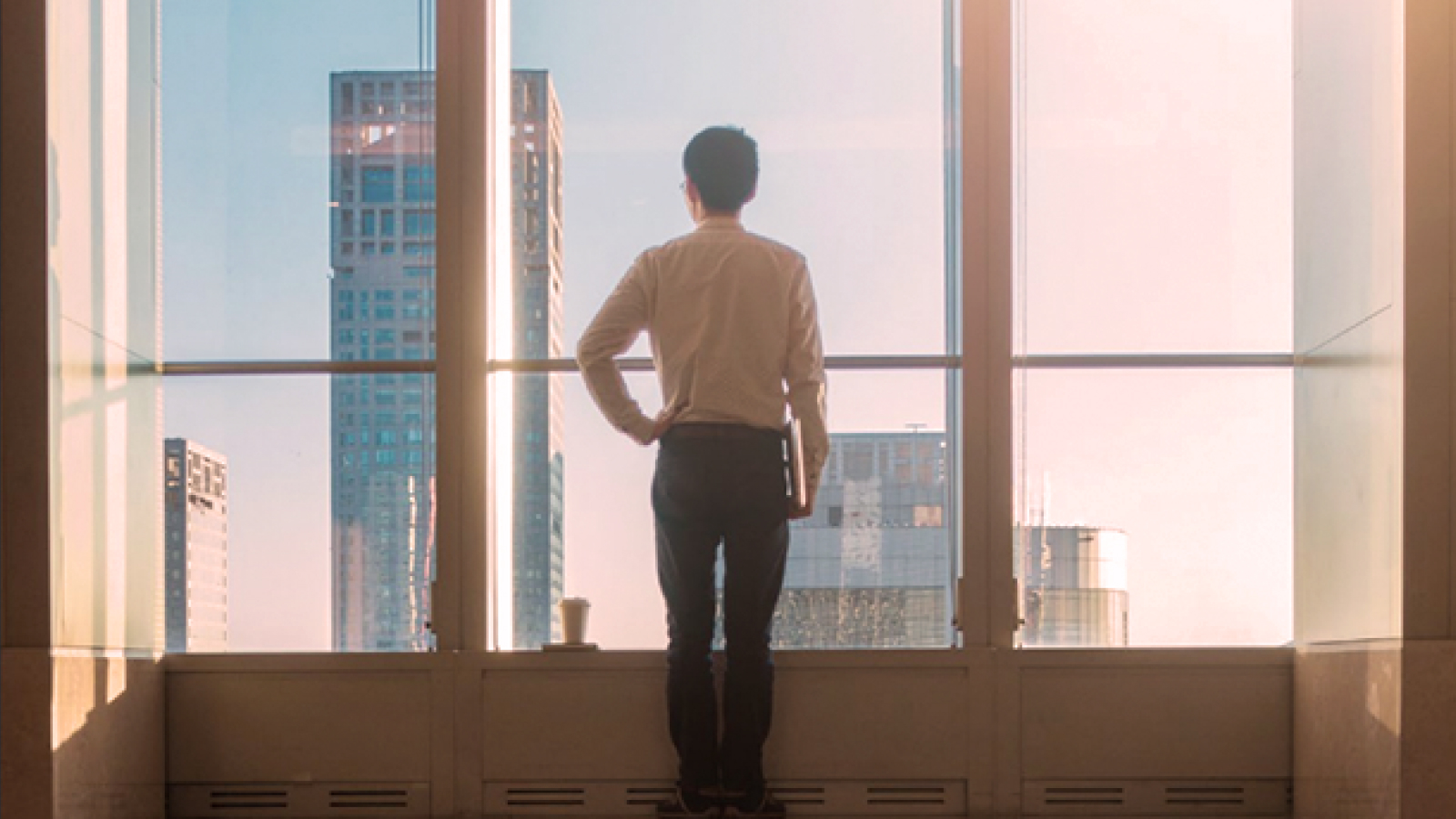 A man stands in front of large floor-to-ceiling windows, overlooking a cityscape with tall buildings at sunrise, his reflection visible on the polished floor.