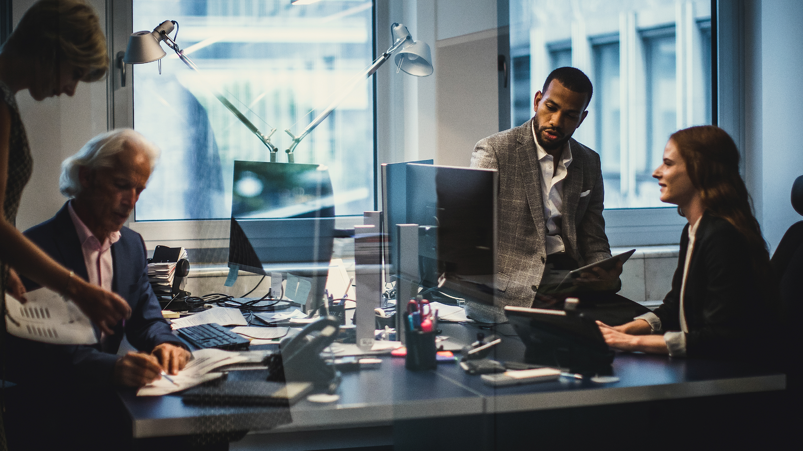 Three professionals in a modern office collaborating around computer monitors and documents, with reflections on glass walls creating a dynamic workspace atmosphere.