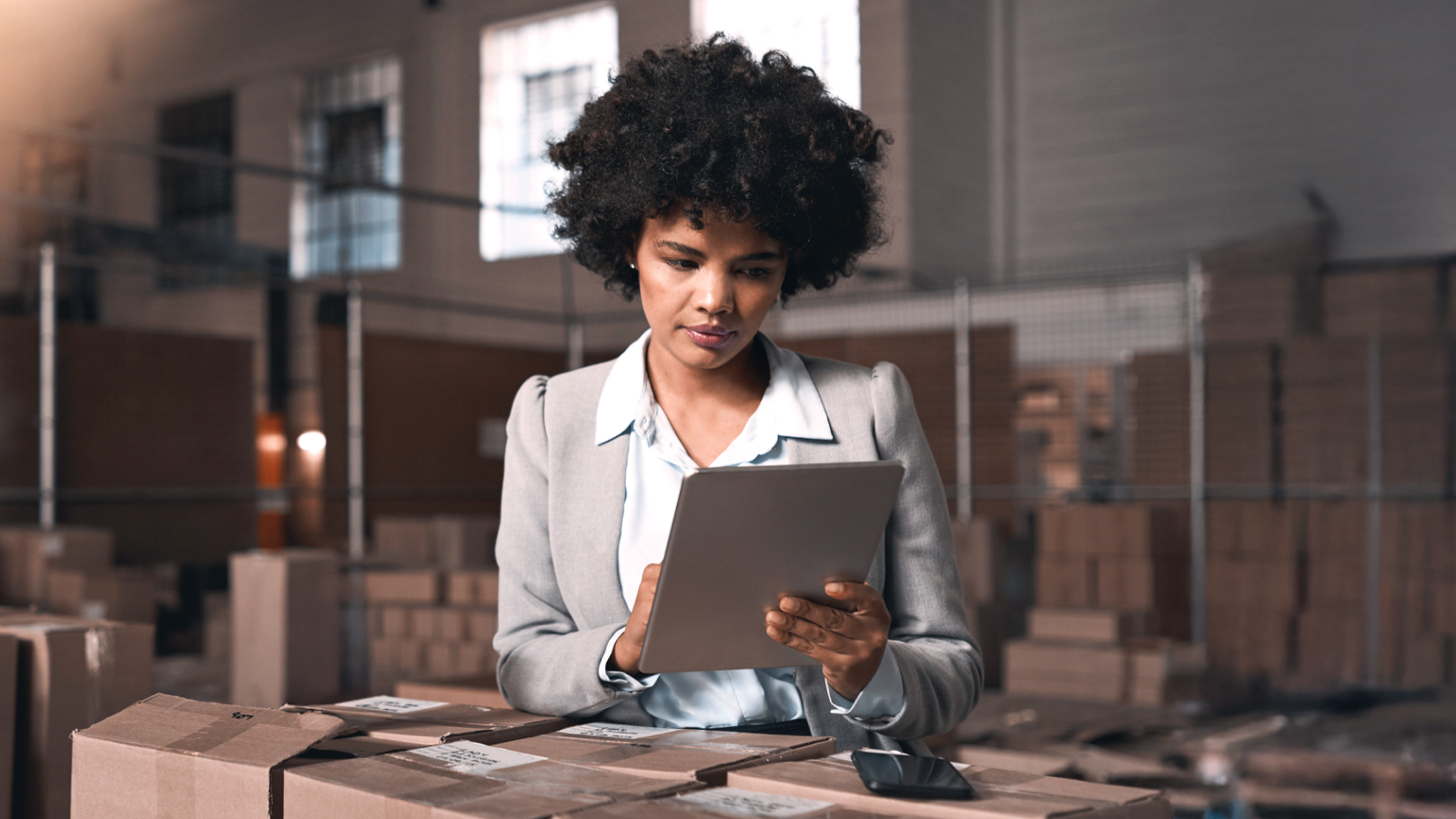 A woman in a warehouse setting focused on a tablet while surrounded by cardboard boxes.