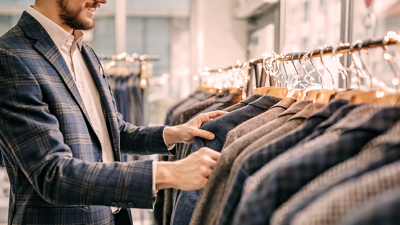 Close-up of a man in a blue plaid blazer examining coats on hangers in a well-lit retail clothing store decorated with warm string lights.