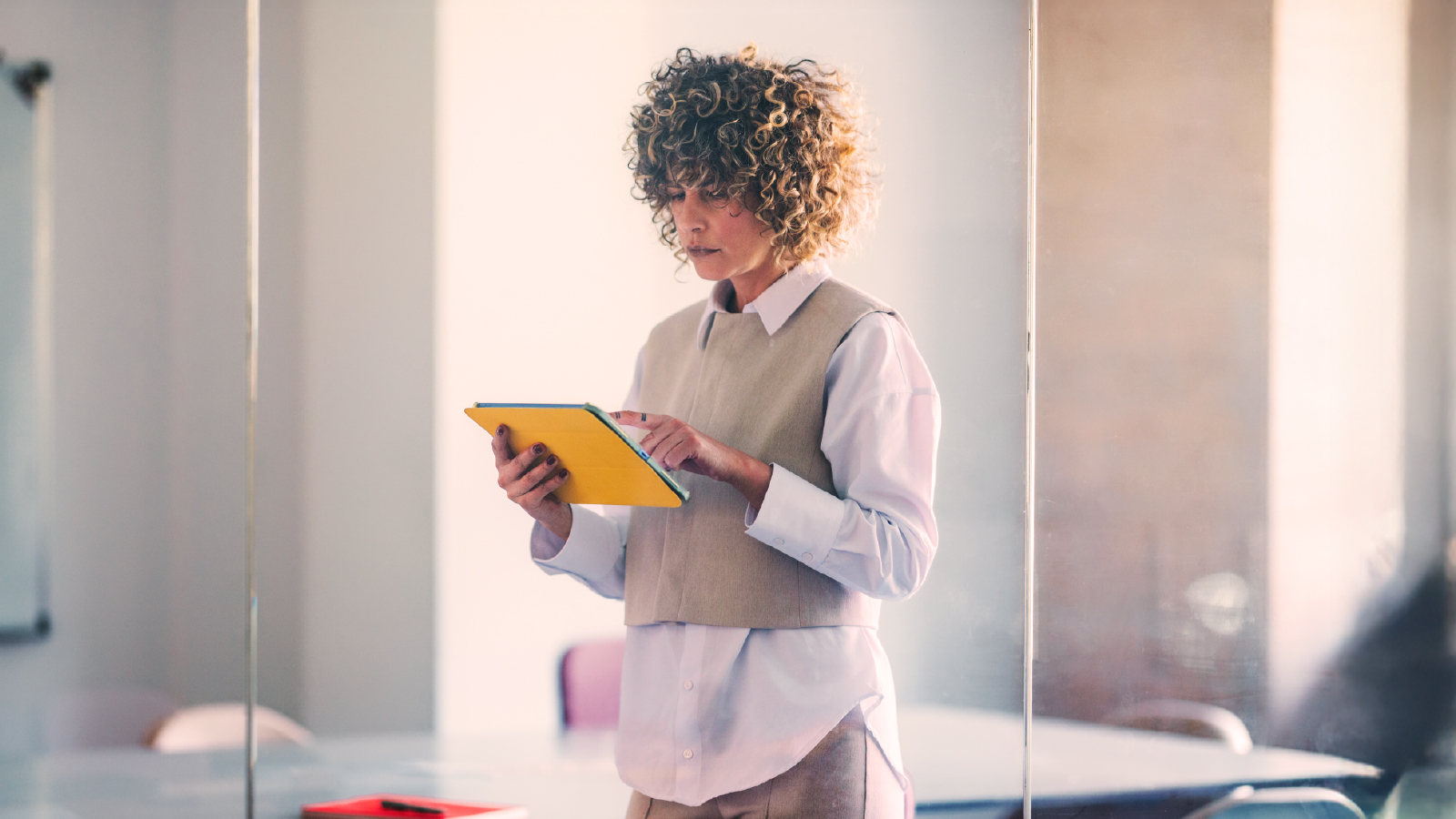 A woman standing in a bright office, holding and using a yellow tablet while reviewing information.