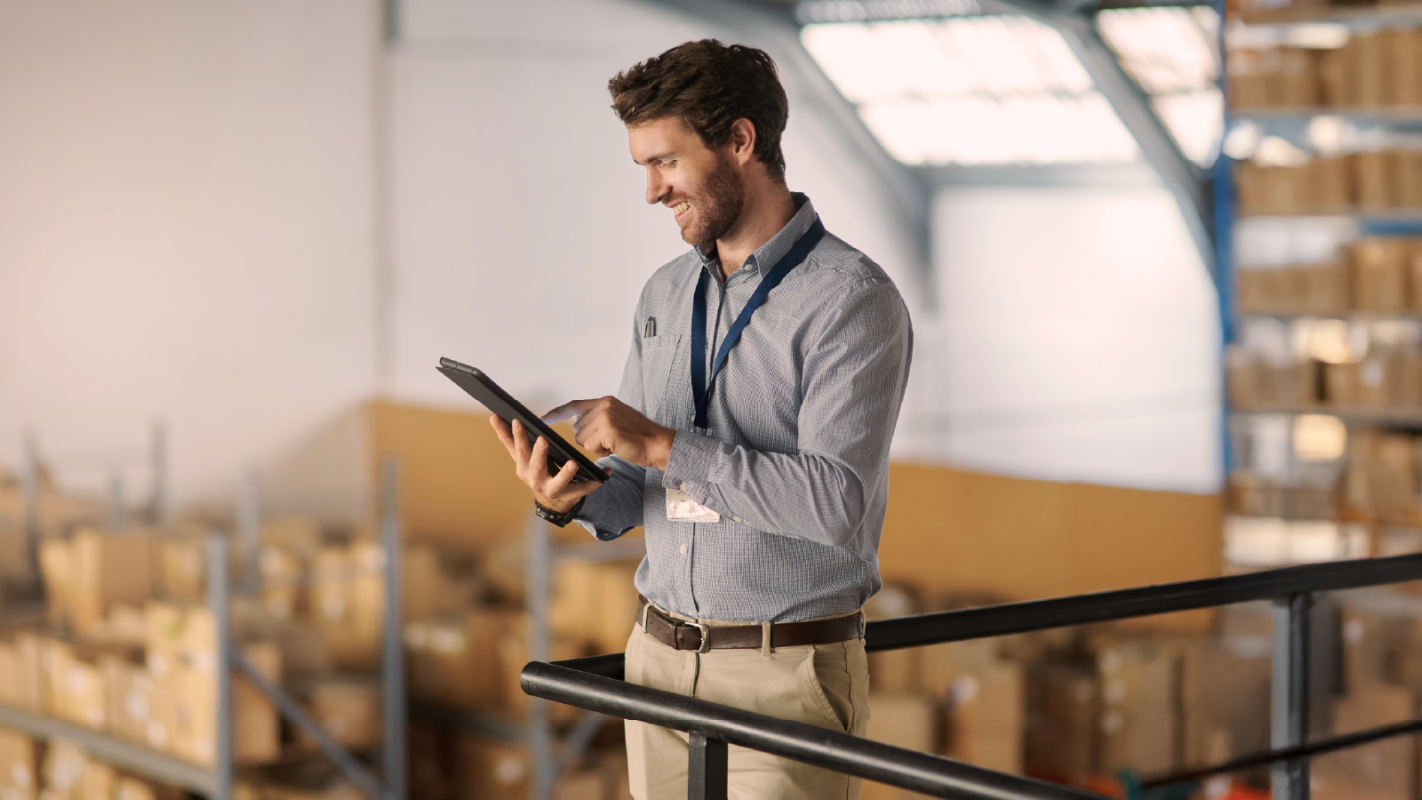 A man in a light blue shirt and lanyard standing on a platform in a warehouse, smiling as he uses a tablet, with shelves of boxes in the background.