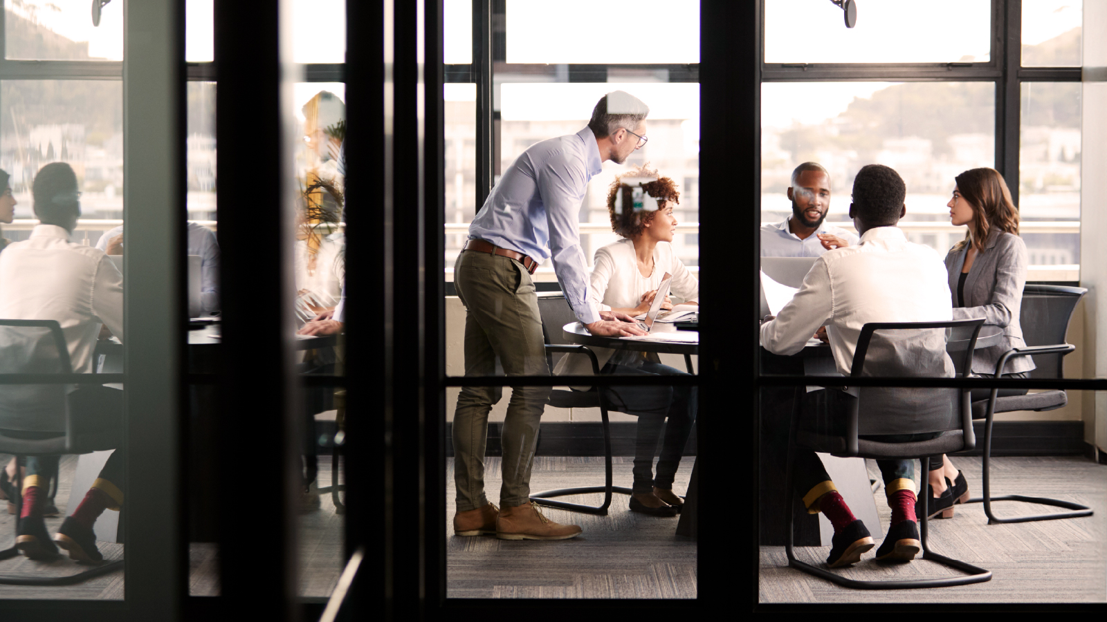Five professionals in a glass-walled meeting room engaged in discussion around a table, with one person standing and leaning in to contribute.
