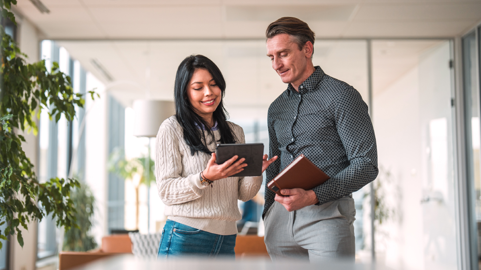 Two colleagues smiling and discussing something on a tablet in a modern office with bright natural light.