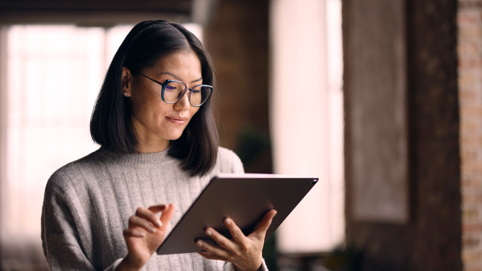 A woman in glasses and a grey sweater uses a digital tablet while sitting indoors with soft natural light.
