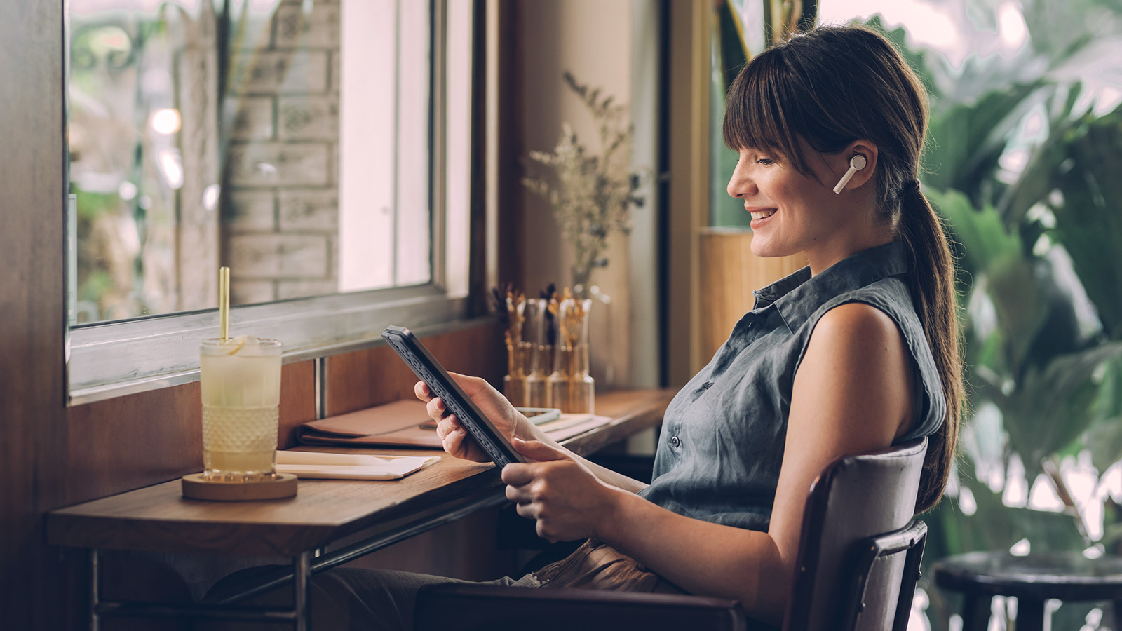 Person sitting at a café counter with a drink, wearing wireless earbuds and smiling while viewing a tablet near a window surrounded by plants.