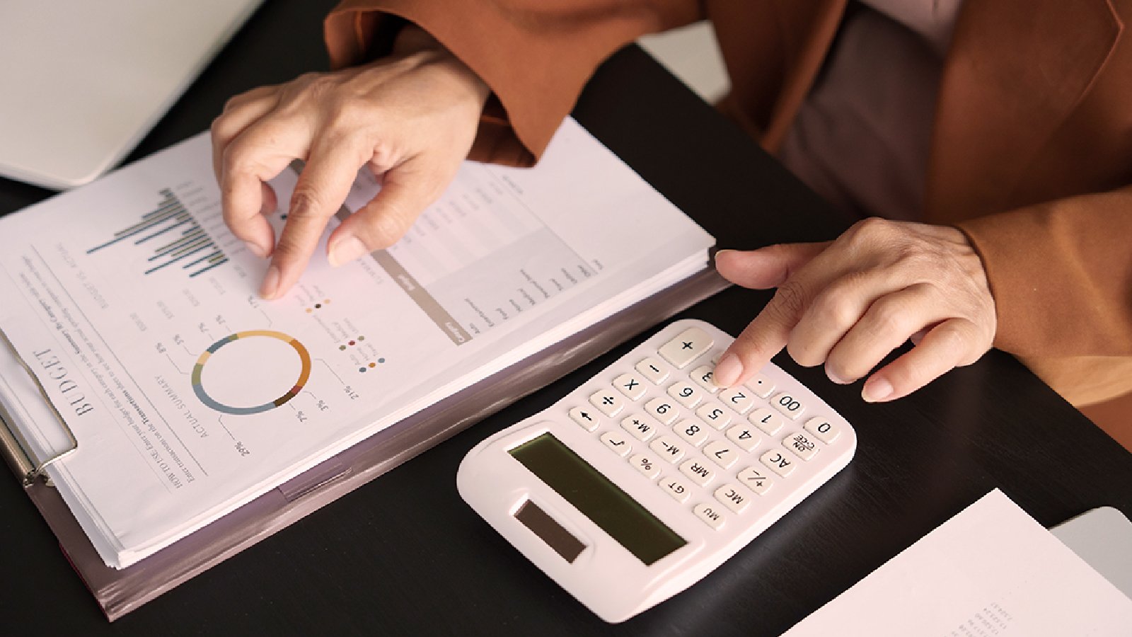 A close-up of a person's hands working on financial documents, pointing at a budget report with charts and graphs while using a white calculator on a dark desk.
