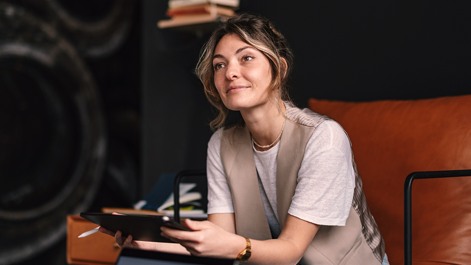 A professional woman sitting on an orange sofa in a modern workspace, holding a tablet and appearing thoughtful.