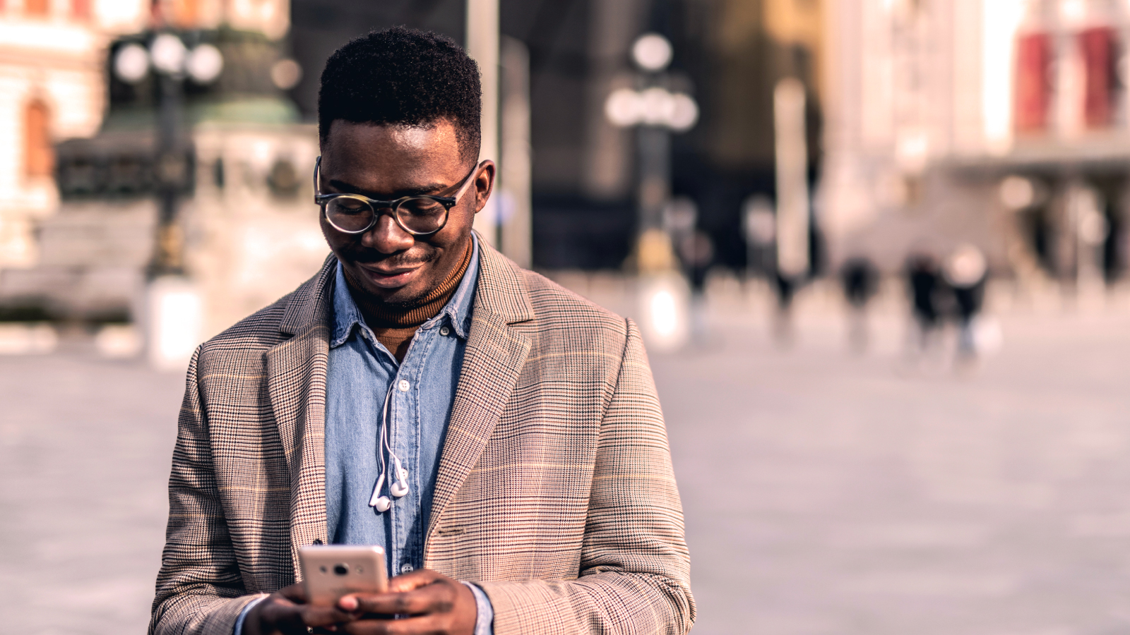A man in a checkered blazer and glasses smiles while looking at his smartphone outdoors in a city square.