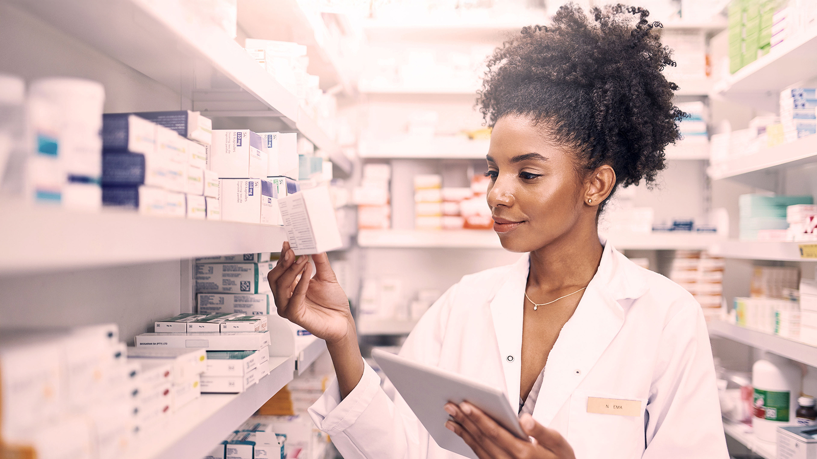 Pharmacist in a white coat examining a medicine box while holding a tablet, surrounded by shelves filled with pharmaceutical products.