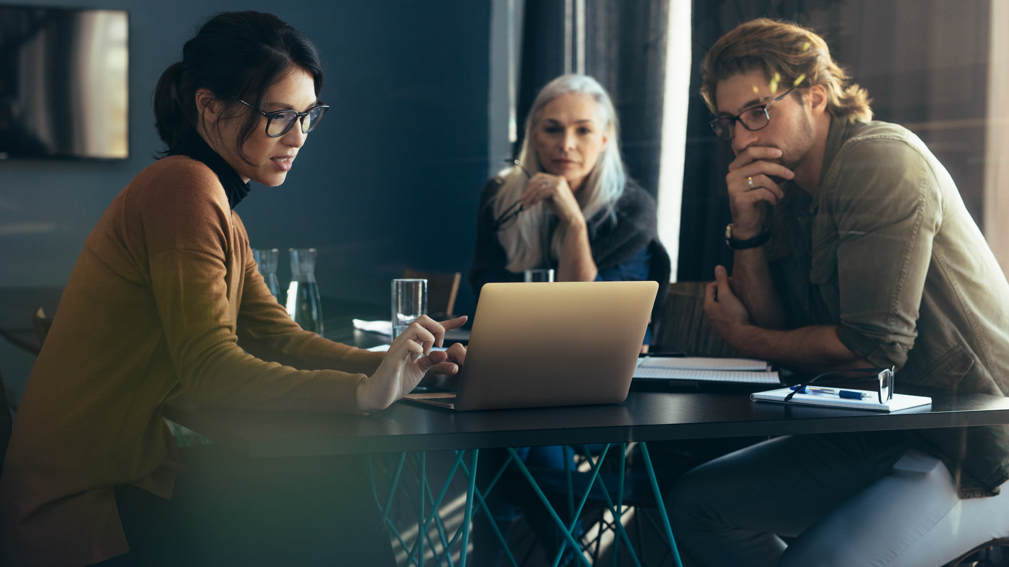 Three colleagues sit around a table in an office, reviewing information on a laptop and discussing work-related matters, with notebooks, pens, and glasses of water nearby.