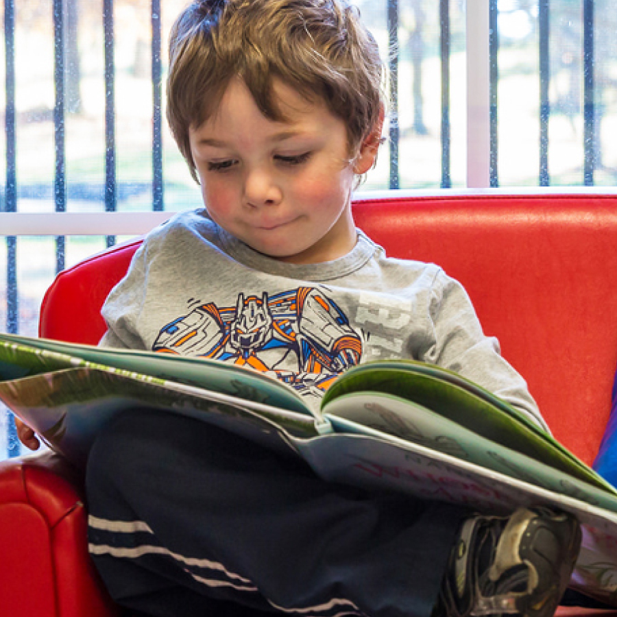 A young boy in a red chair, deeply focused on a book, illustrating a peaceful moment of reading and learning.