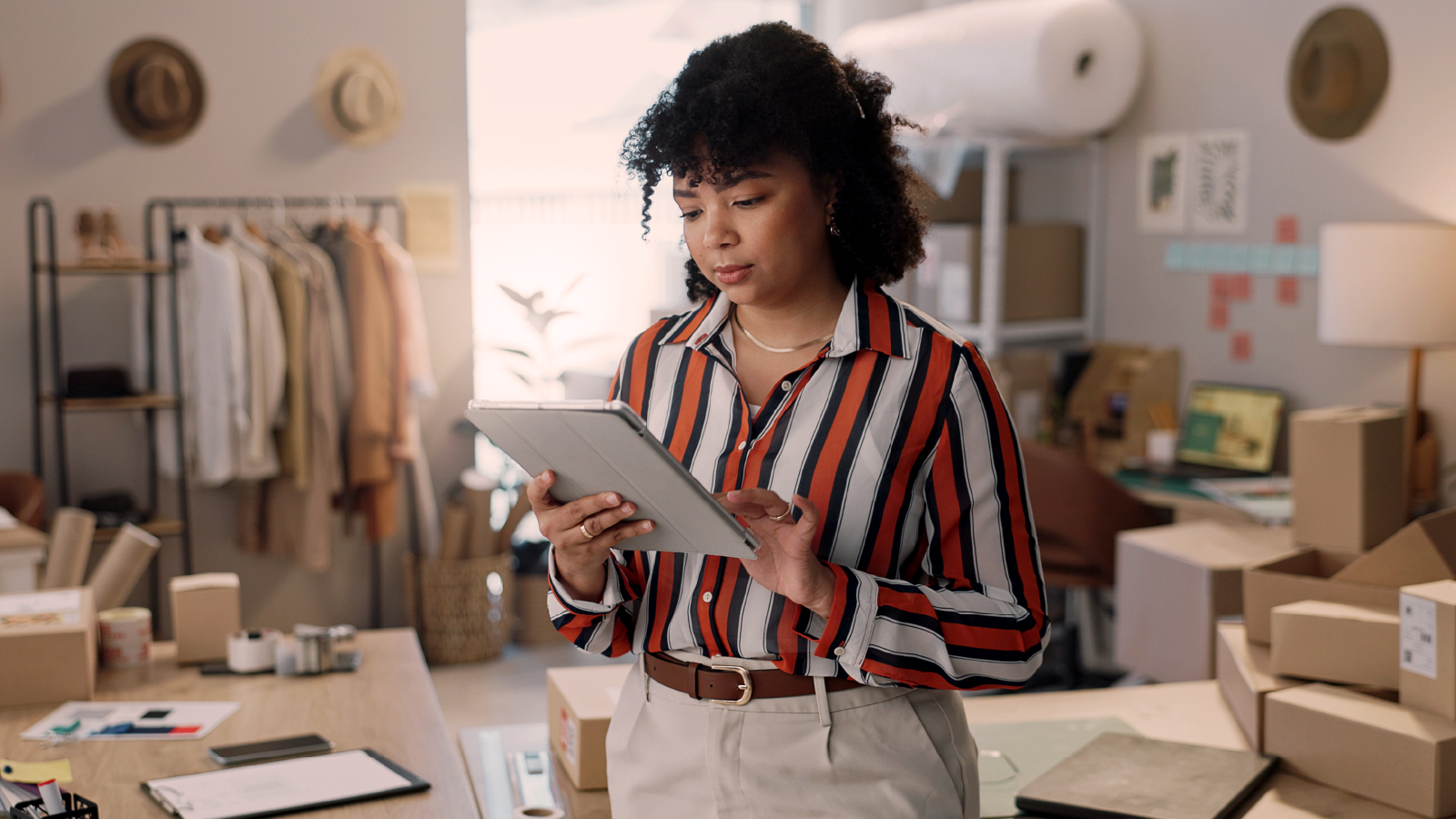 A woman in a striped shirt holding a tablet and reviewing information in a retail workspace with clothing racks and packaging materials in the background.