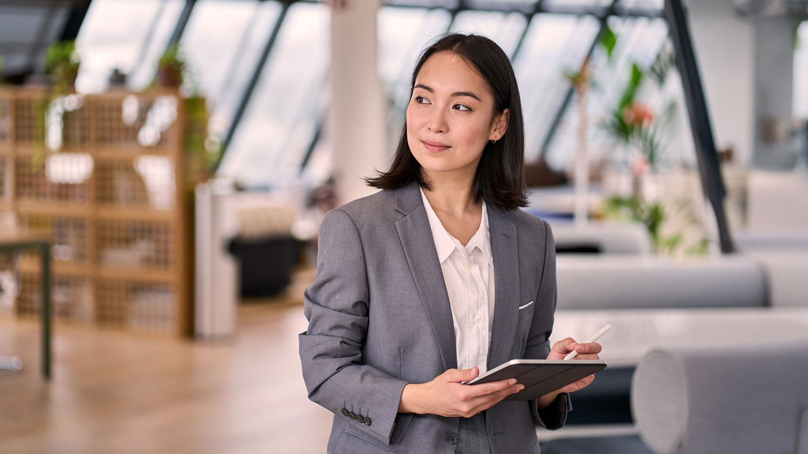 A business professional in a gray suit holding a tablet and stylus, standing in a modern office with large windows and a background featuring greenery and office furniture.