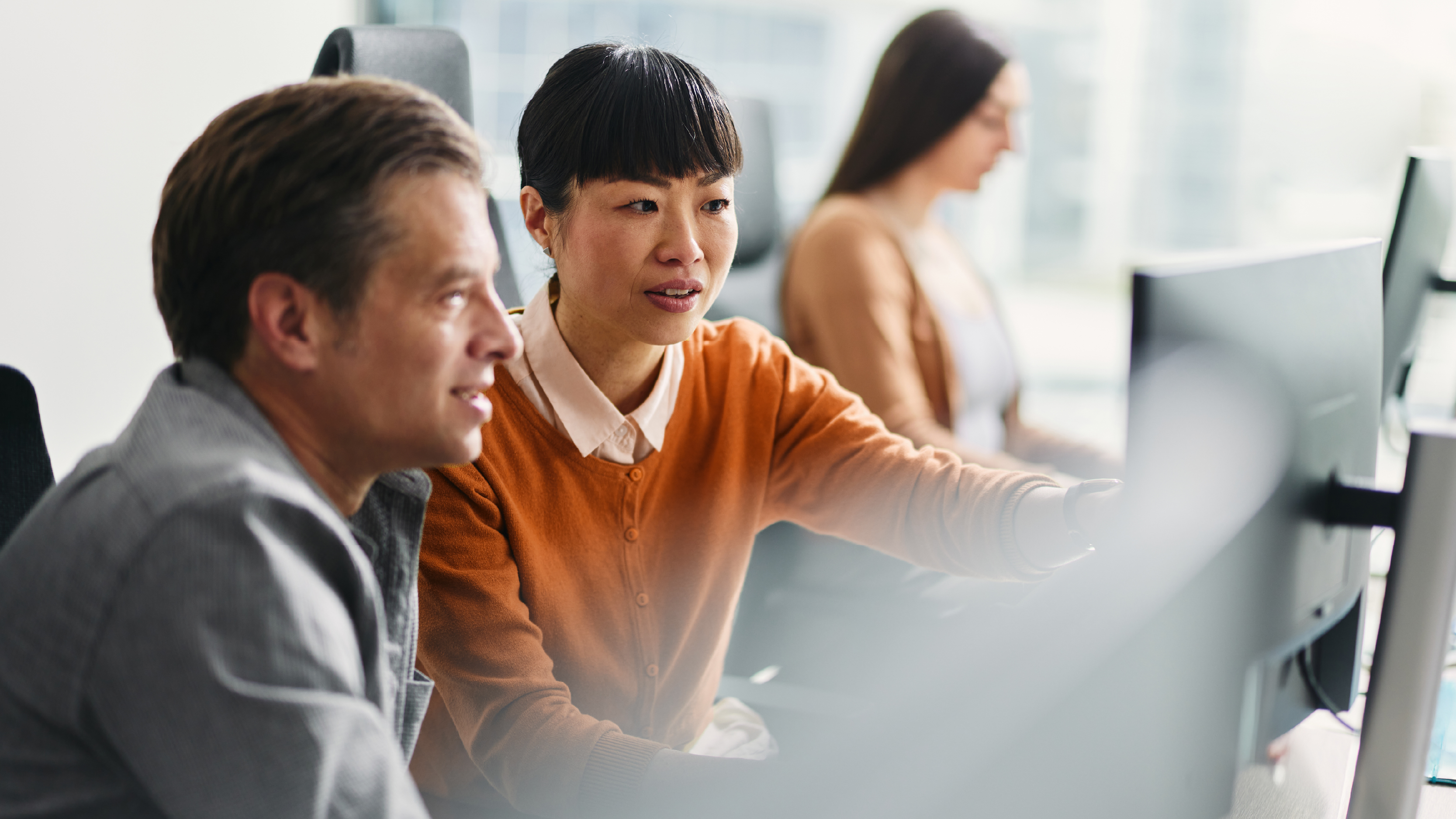 Two professionals focused on a desktop monitor in a bright office space, discussing data or visuals, with a third colleague working in the background.