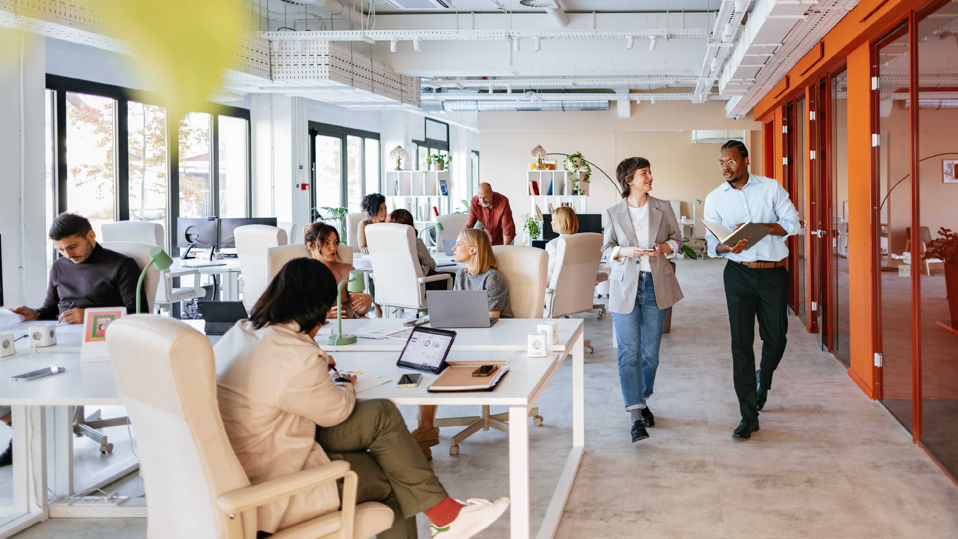 Modern open-plan office with professionals working at desks and in discussion. Two colleagues walk together in conversation near a hallway with glass and orange-framed walls.