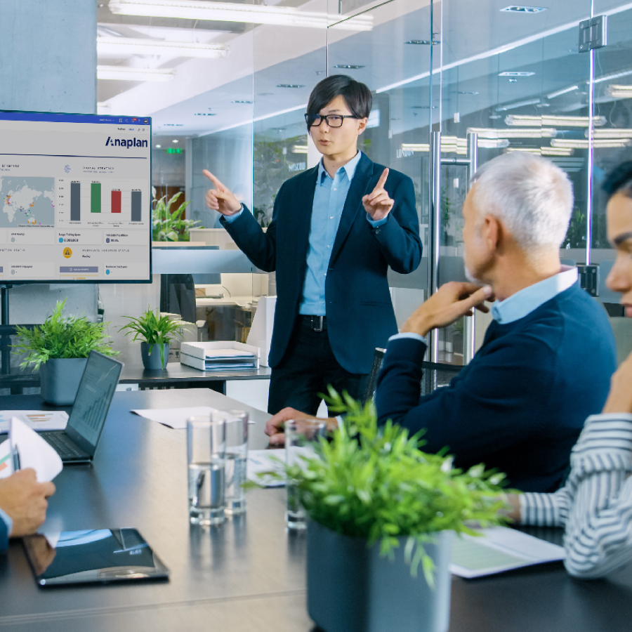 A man presents to a group of people in a meeting room, engaged in discussion and taking notes.