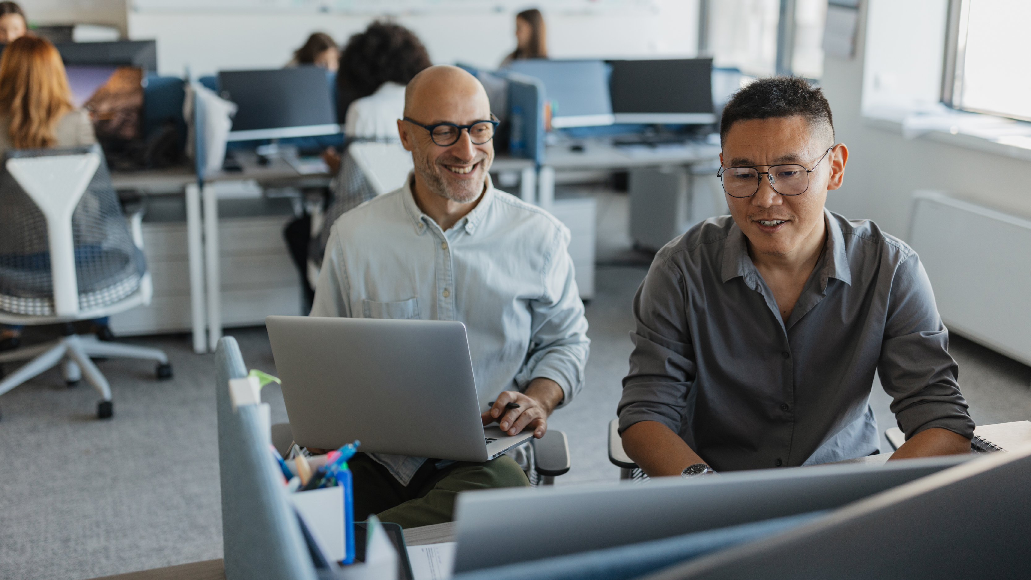 Two male coworkers smiling and working together in a collaborative office space, with laptops and desktop monitors on a shared desk.